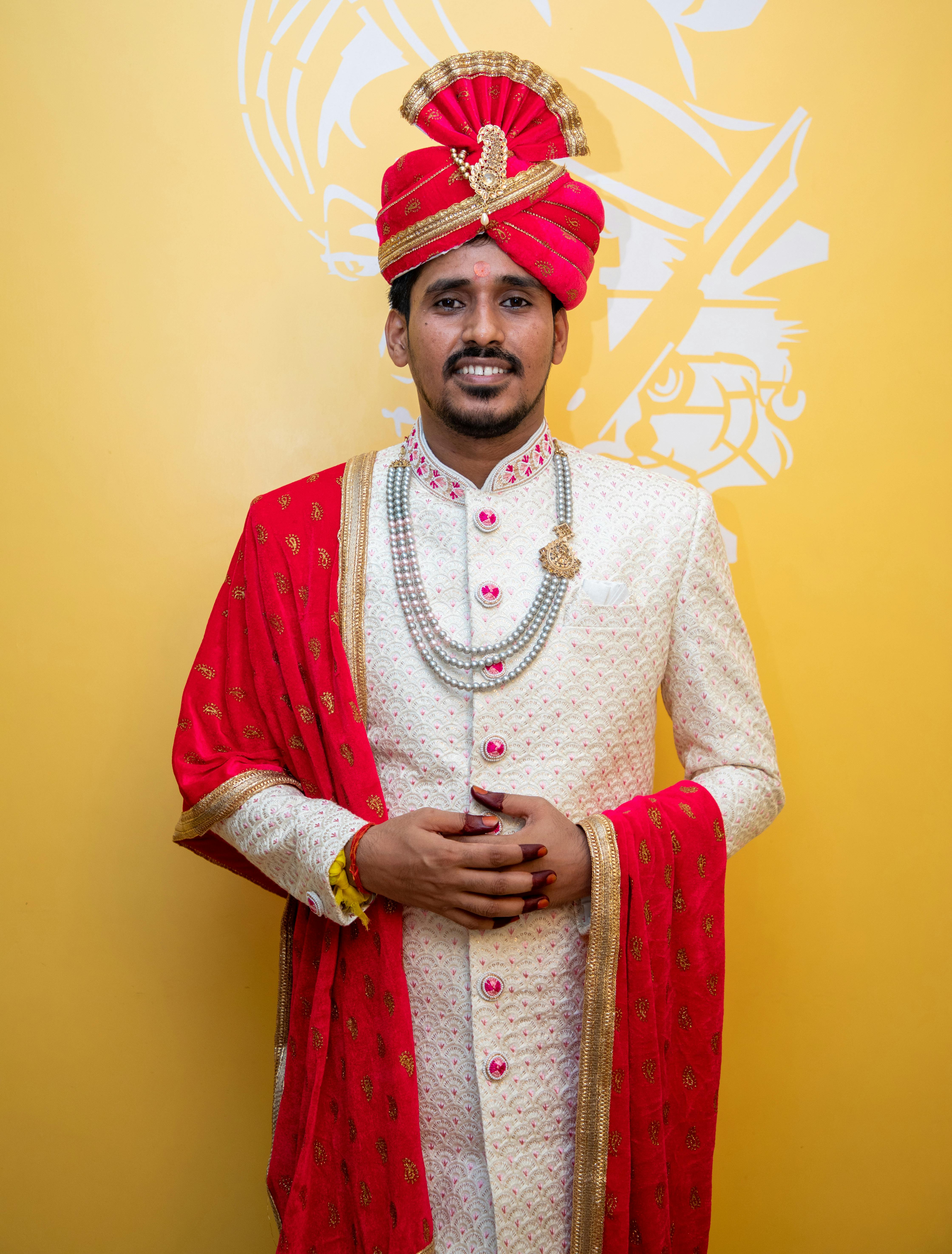 Indian groom wearing traditional wedding attire with vibrant red turban and jewelry.