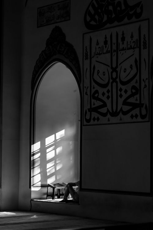 Free Black and white image of a man sitting in a mosque, capturing the play of light and Islamic calligraphy. Stock Photo