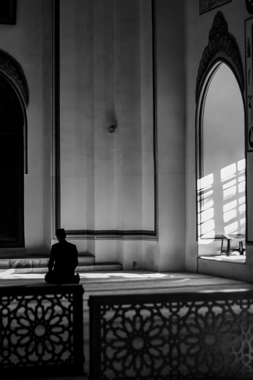 Free Black and white image of a man in prayer at a mosque, enveloped in sunbeams and Islamic architecture. Stock Photo