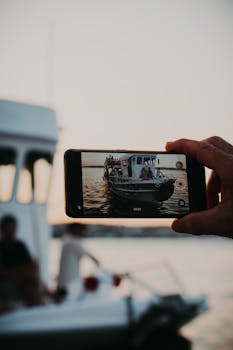 A hand holding a smartphone capturing a motorboat scene in Ayvalık, Türkiye at sunset.
