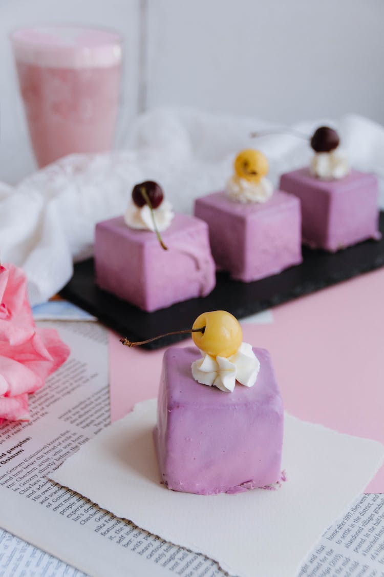 Lavender Colored Glazed Petit Four Cakes With Yellow And Red Cherries And A Glass Of Fruit Milkshake