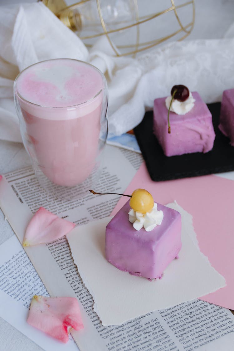 Glass Of Pink Milkshake With Lavender Colored Petit Four Cakes With Cherry And Cream Topping