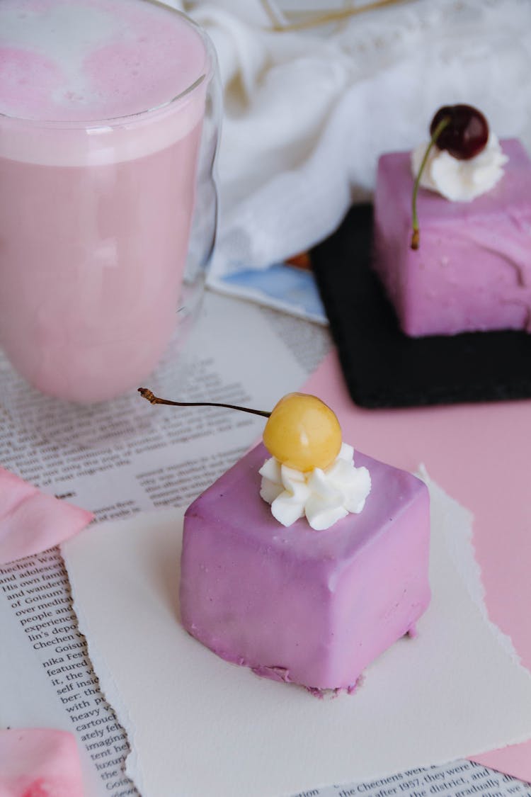 Lilac Colored Petit Four Cake With Yellow Cherry And A Glass Of Fruit Milkshake