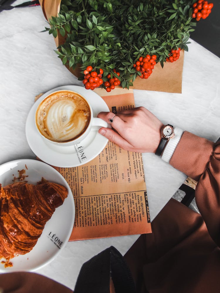 Person Sitting At A Table With A Cup Of Coffee And Croissant