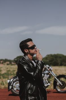 A stylish man in a leather jacket smokes near a motorcycle outdoors.