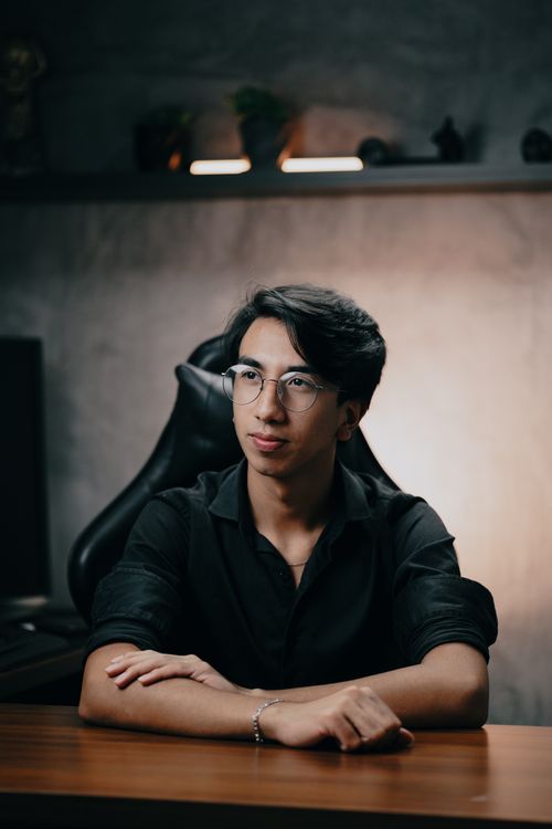 Free Young man wearing glasses sitting at a modern desk in casual office setting. Stock Photo