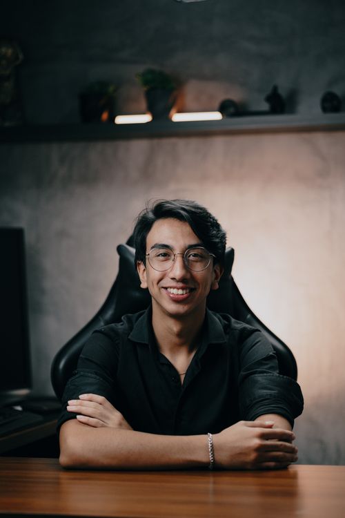 Free A young man with glasses sitting confidently at a desk, smiling in a modern office environment. Stock Photo