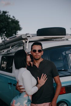 A young couple embraces while posing in front of a vintage van, outdoors.