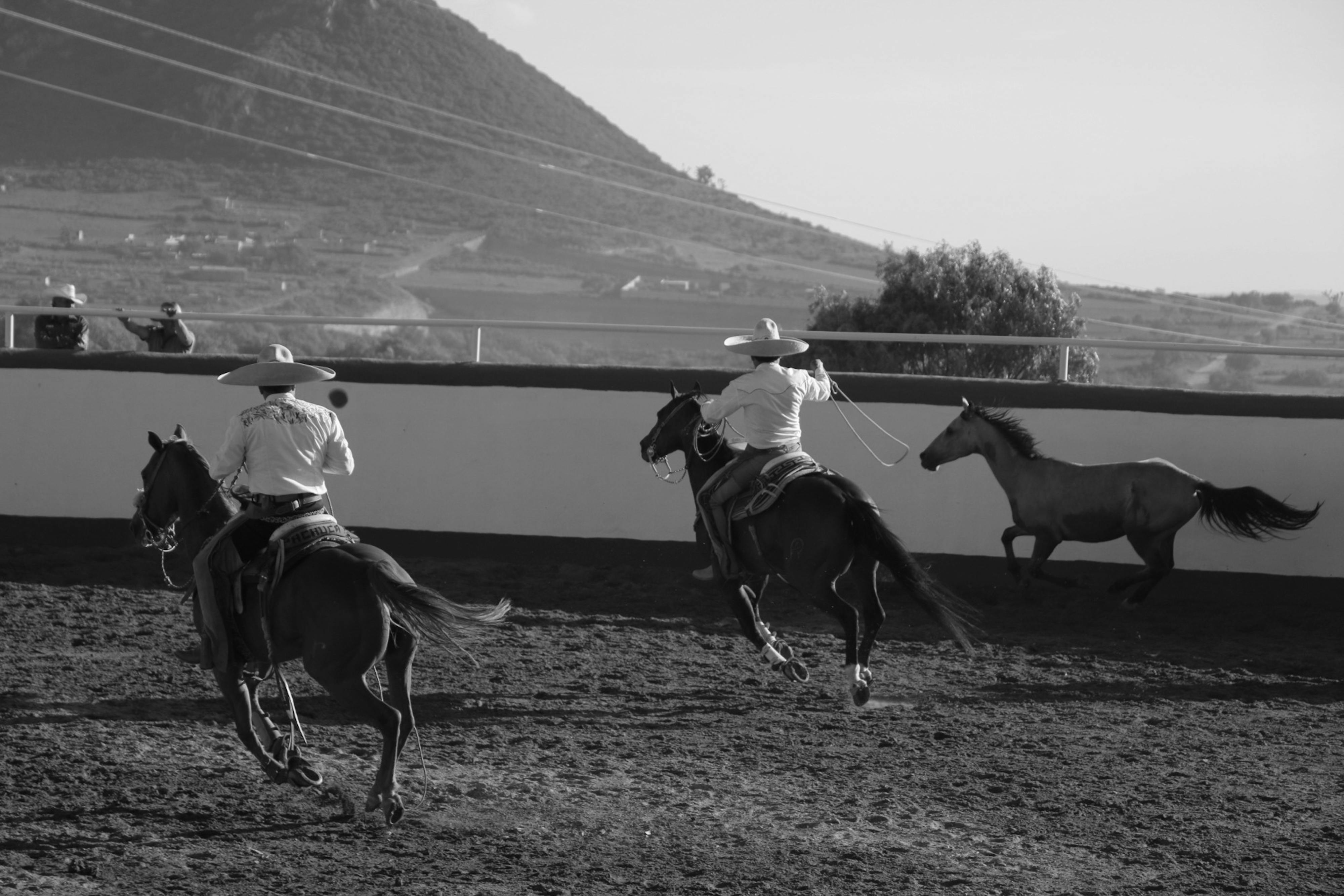 Black and white photo of horseback riders in traditional attire at outdoor rodeo.