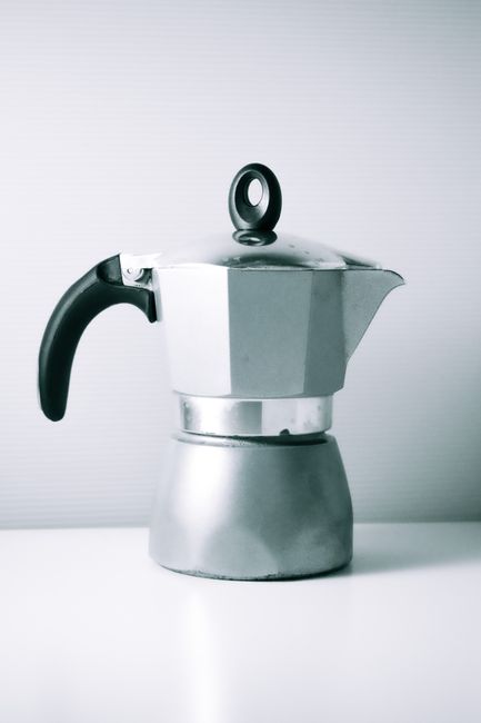 A stylish silver moka pot coffee maker with a black handle resting on a clean white surface in a minimalist setting.