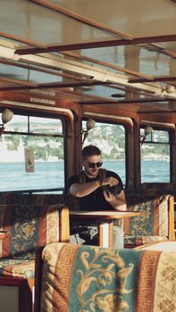 A young man using a camera while enjoying a ferry ride in Istanbul with a scenic view in the background.