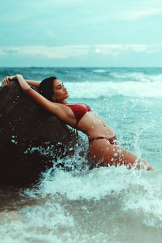 Elegant woman in a red bikini poses on a rock by the ocean, waves crashing around her.