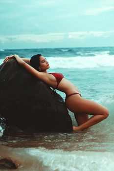 Woman posing in a red bikini on a rocky shore with waves crashing around her.