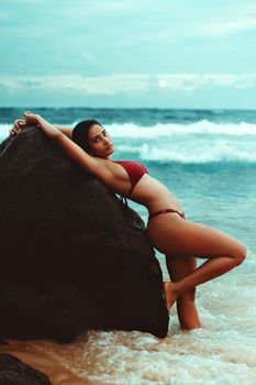 A woman poses in a red bikini beside ocean rocks, capturing the essence of beach fashion.