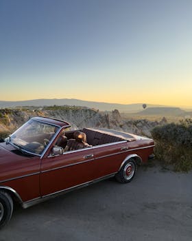 Classic red car set against Cappadocia's unique landscape during sunset, featuring a serene balloon-filled sky.