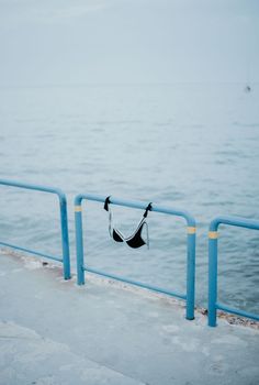 A solitary black bra hangs on a blue railing above a tranquil sea view, suggesting themes of solitude and serenity.