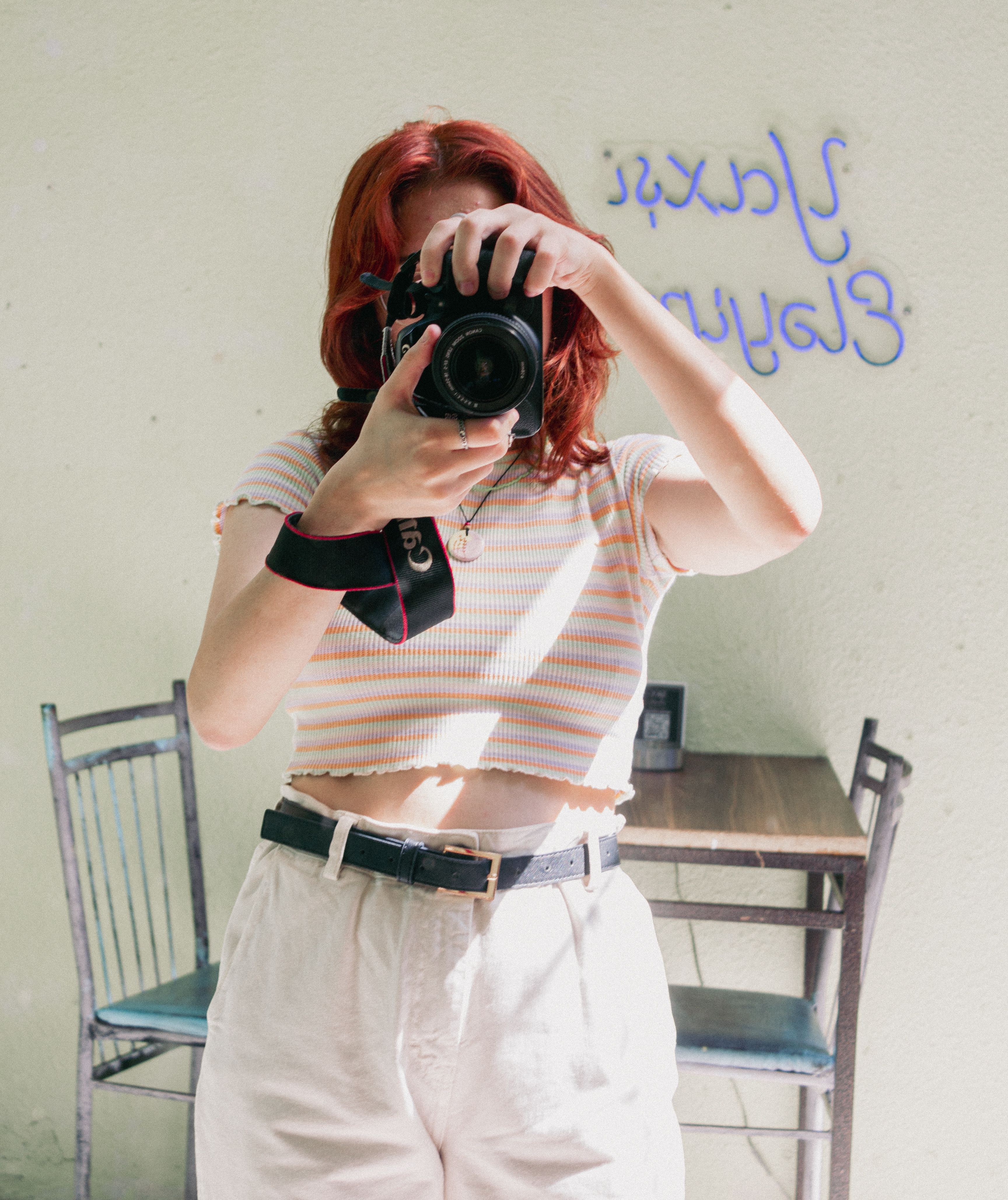 A young woman with red hair takes a selfie in a mirror using a DSLR camera indoors.