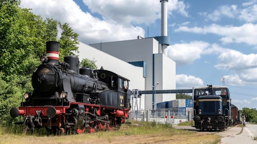 View of Old Locomotive and a Train in front of a Building 