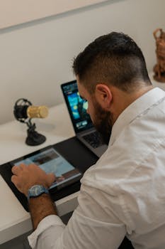 Man in white shirt working at desk with tablet and laptop, engaging with technology.