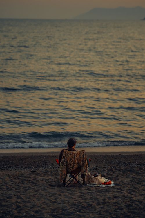 Free A person sits alone on a sandy beach at dusk, enjoying the calm ocean view. Stock Photo