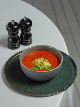 Delicious tomato soup garnished with basil leaves, served on a rustic plate with pepper shakers.