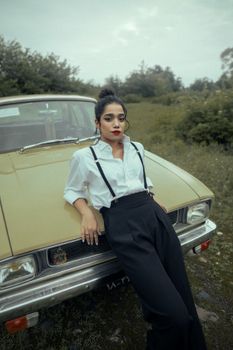 Elegant woman in retro style leaning on a vintage car in a grassy outdoor setting.