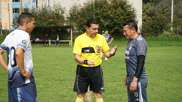 Referee and players in discussion before a soccer match outdoors on a sunny day.