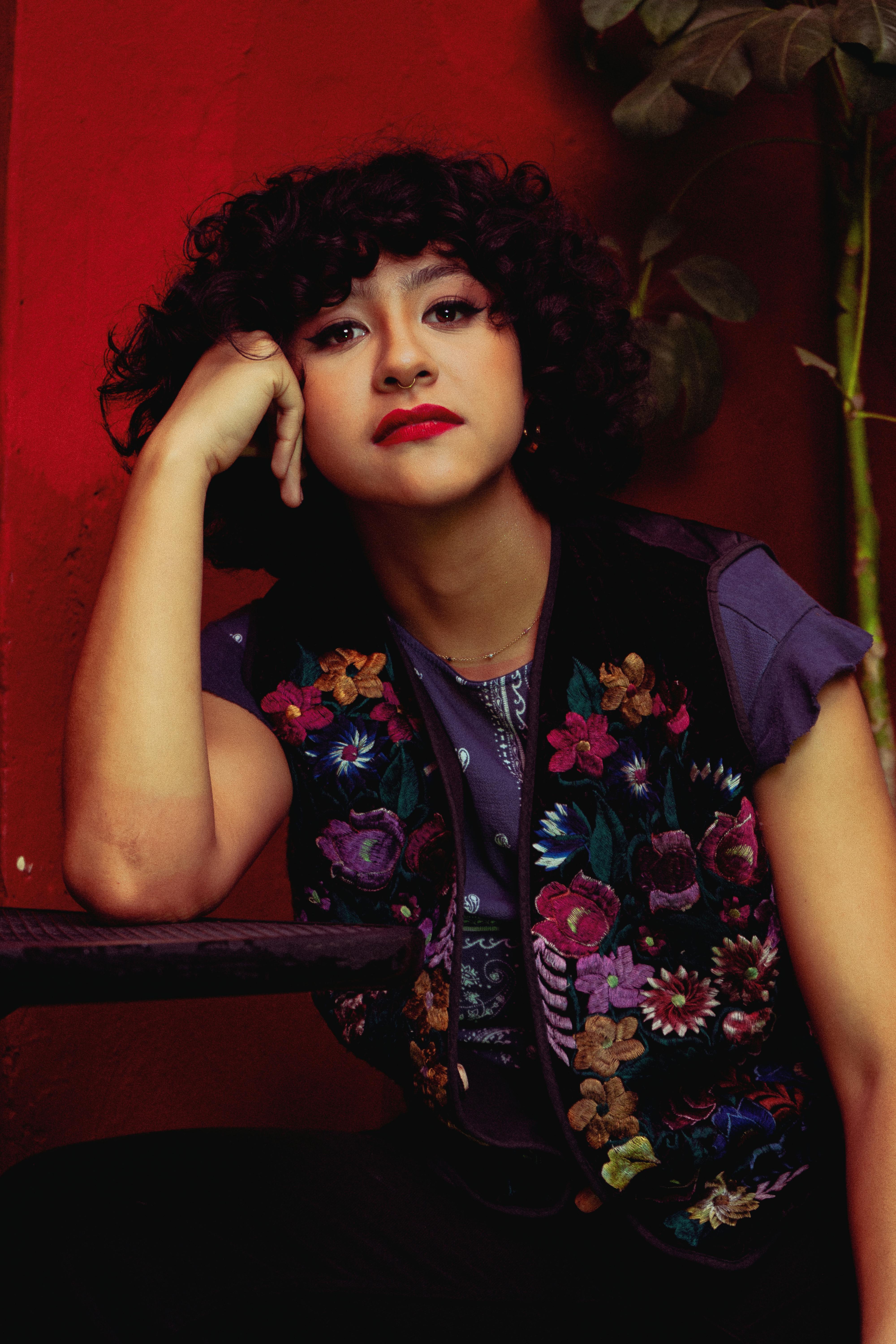 Young woman with curly hair and red lips posing indoors with a floral vest.