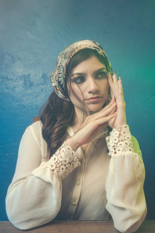 Photo of a Brunette Wearing a Headscarf and a White Blouse, against a Blue Wall