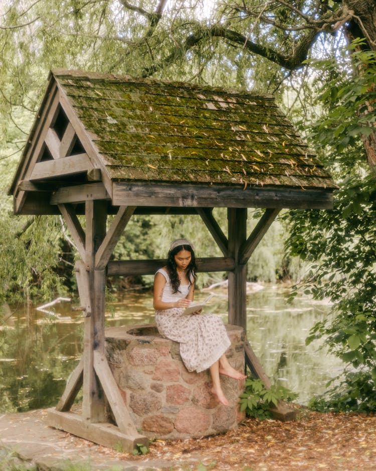 Woman In Dress Sitting On Well In Forest