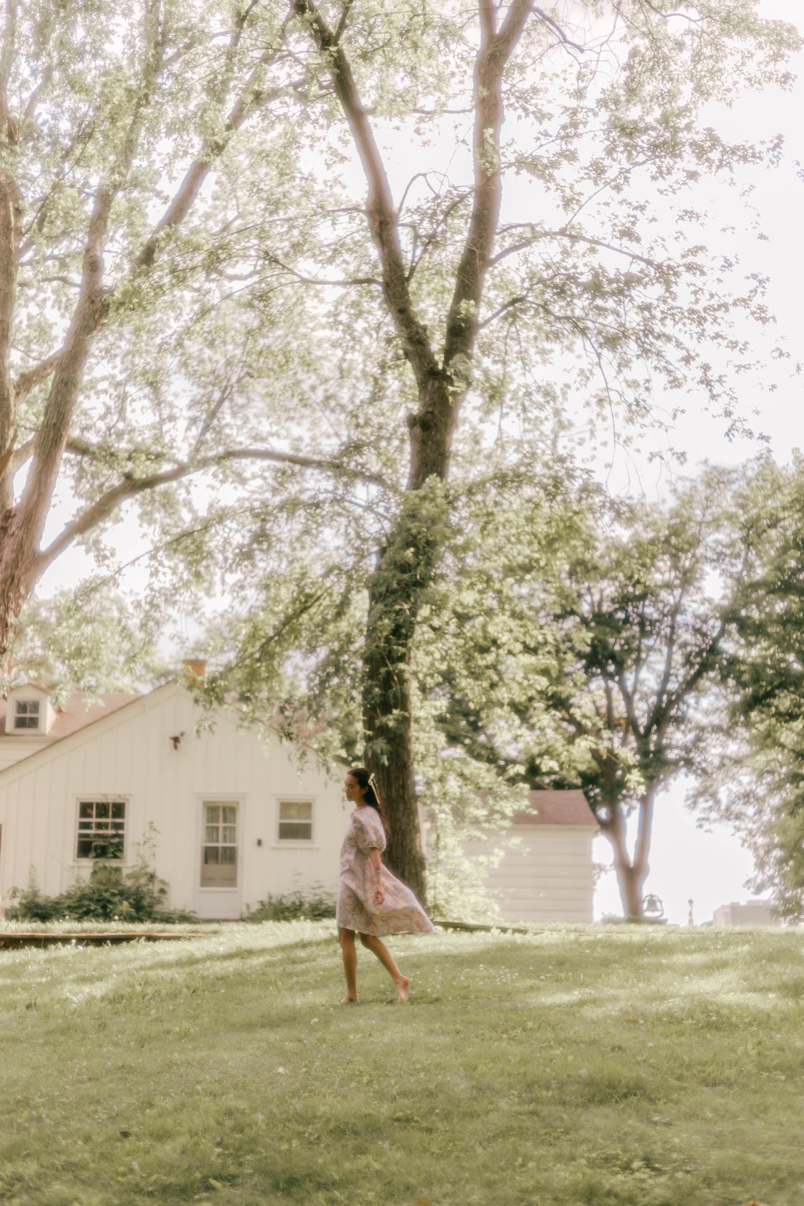 A woman walks gracefully on lush grass near a charming house, under tall trees on a sunny day.