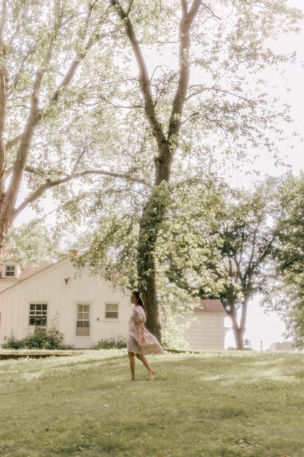A woman walks gracefully on lush grass near a charming house, under tall trees on a sunny day.