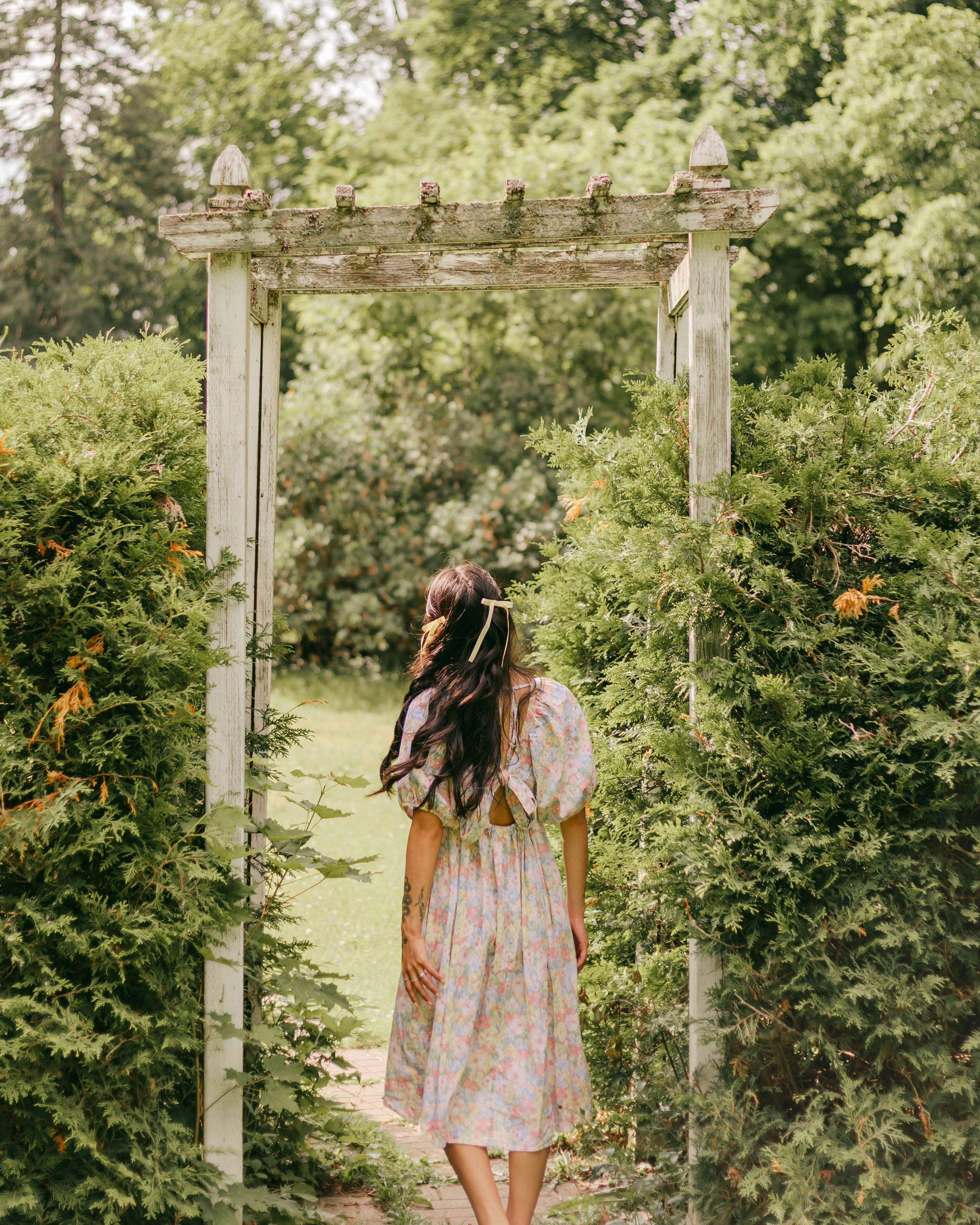 Young woman in a floral dress walking through a garden archway, surrounded by lush greenery.