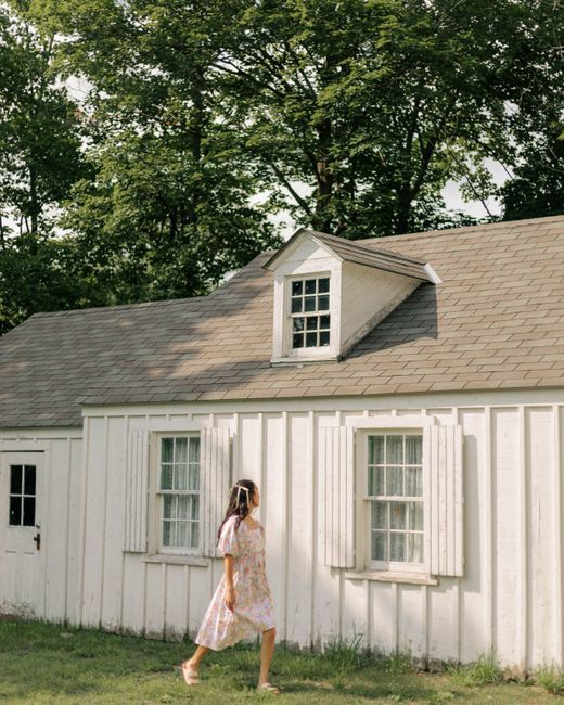 A young woman in a dress walks by a rustic village house during a summer day.