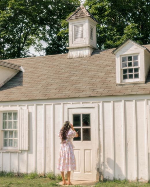 A woman in a floral dress stands by a charming rustic house, capturing a serene summer moment.