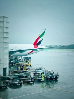 A passenger jet with Emirates livery parked at a rainy airport tarmac with equipment and staff visible.