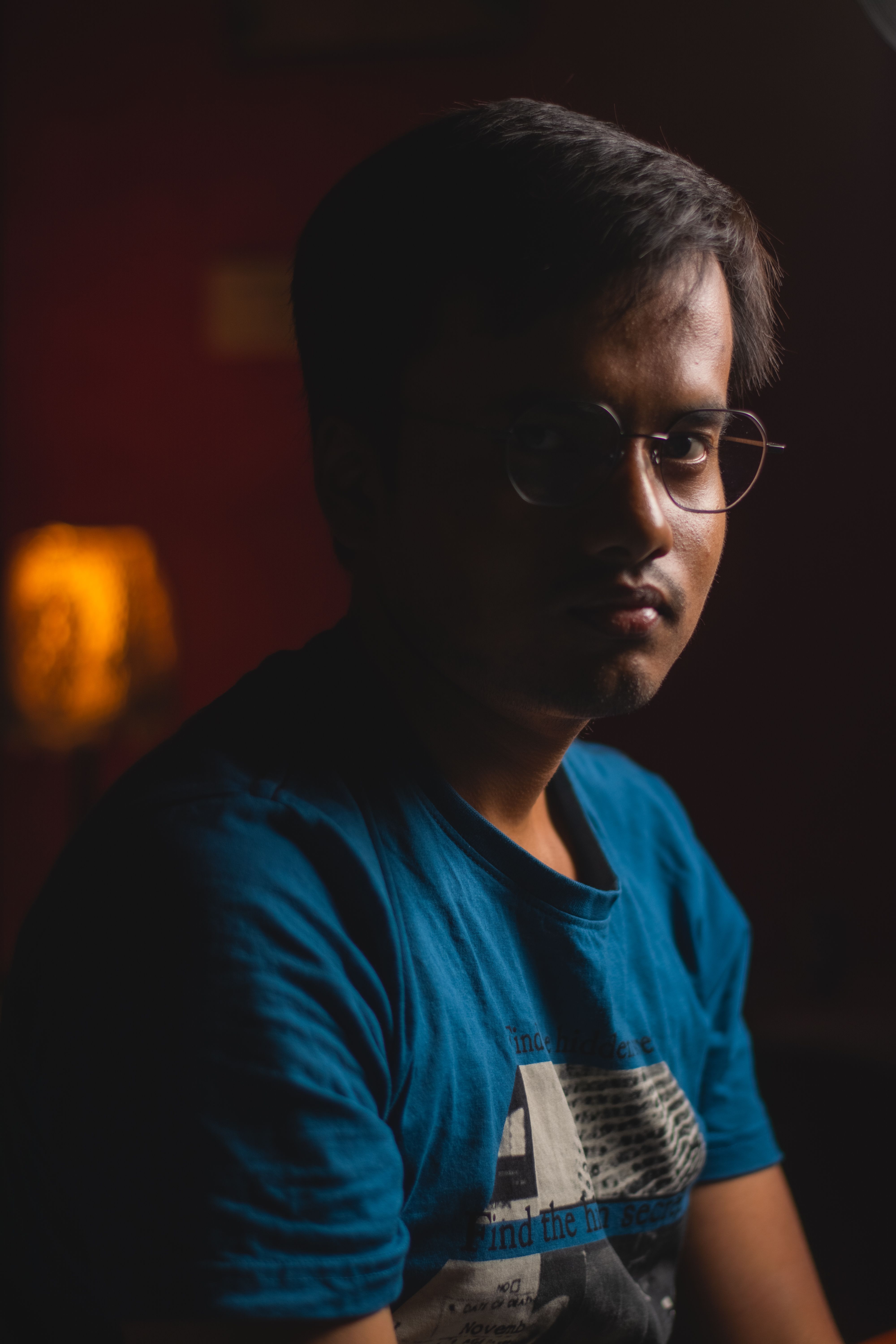 Portrait of a young man in eyeglasses, sitting indoors with a serious expression and dark lighting.
