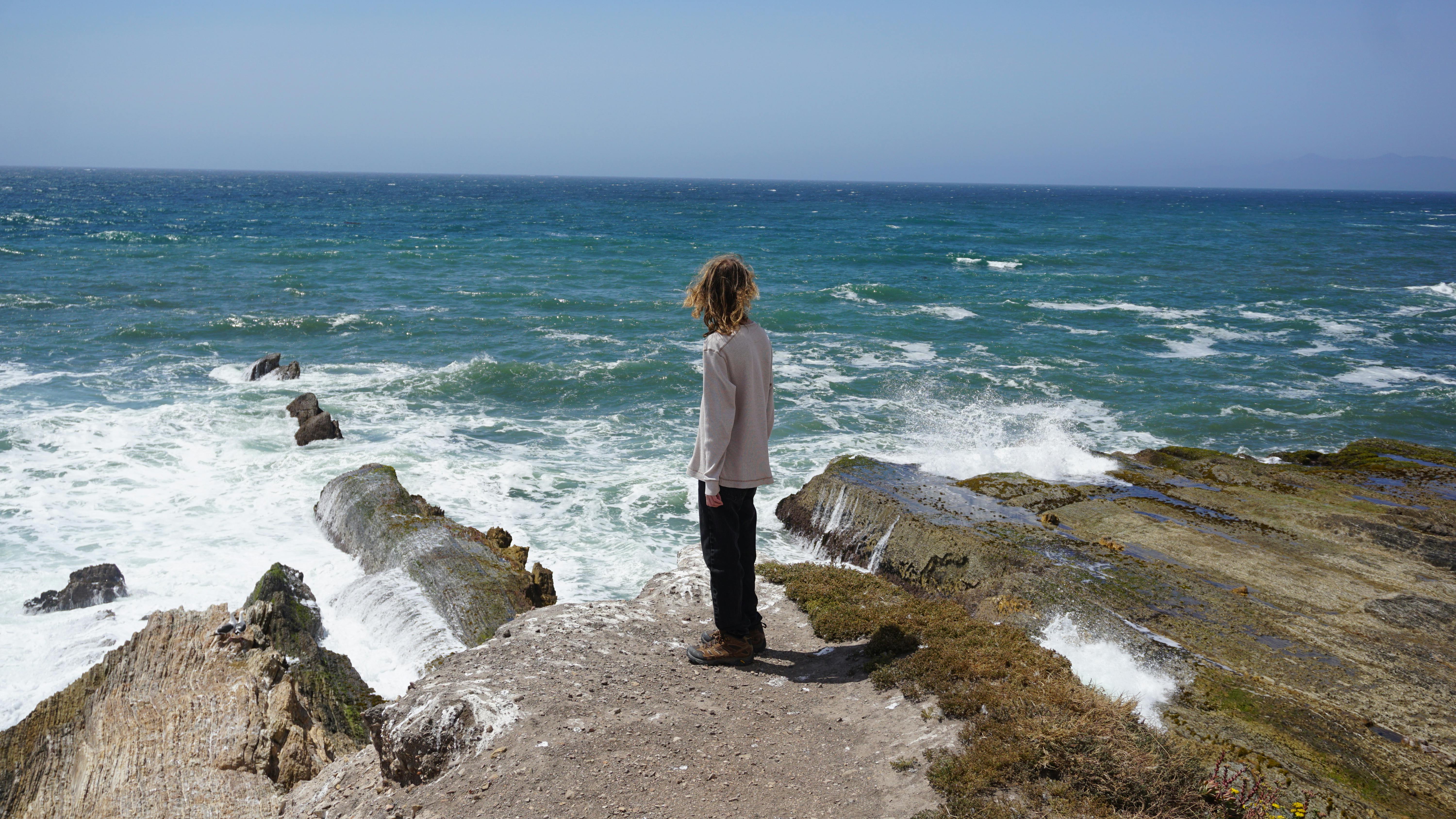 A person stands on a sunlit rocky shore in San Luis Obispo, overlooking the vast sea.