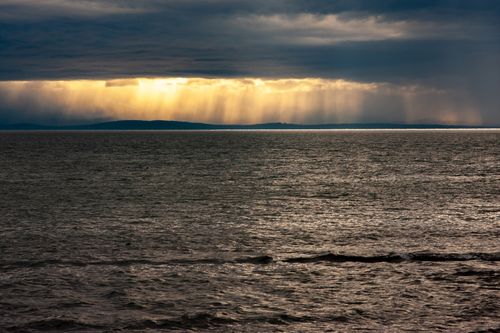 Free Sunbeams break through overcast clouds, casting light over a calm sea at dusk. Stock Photo