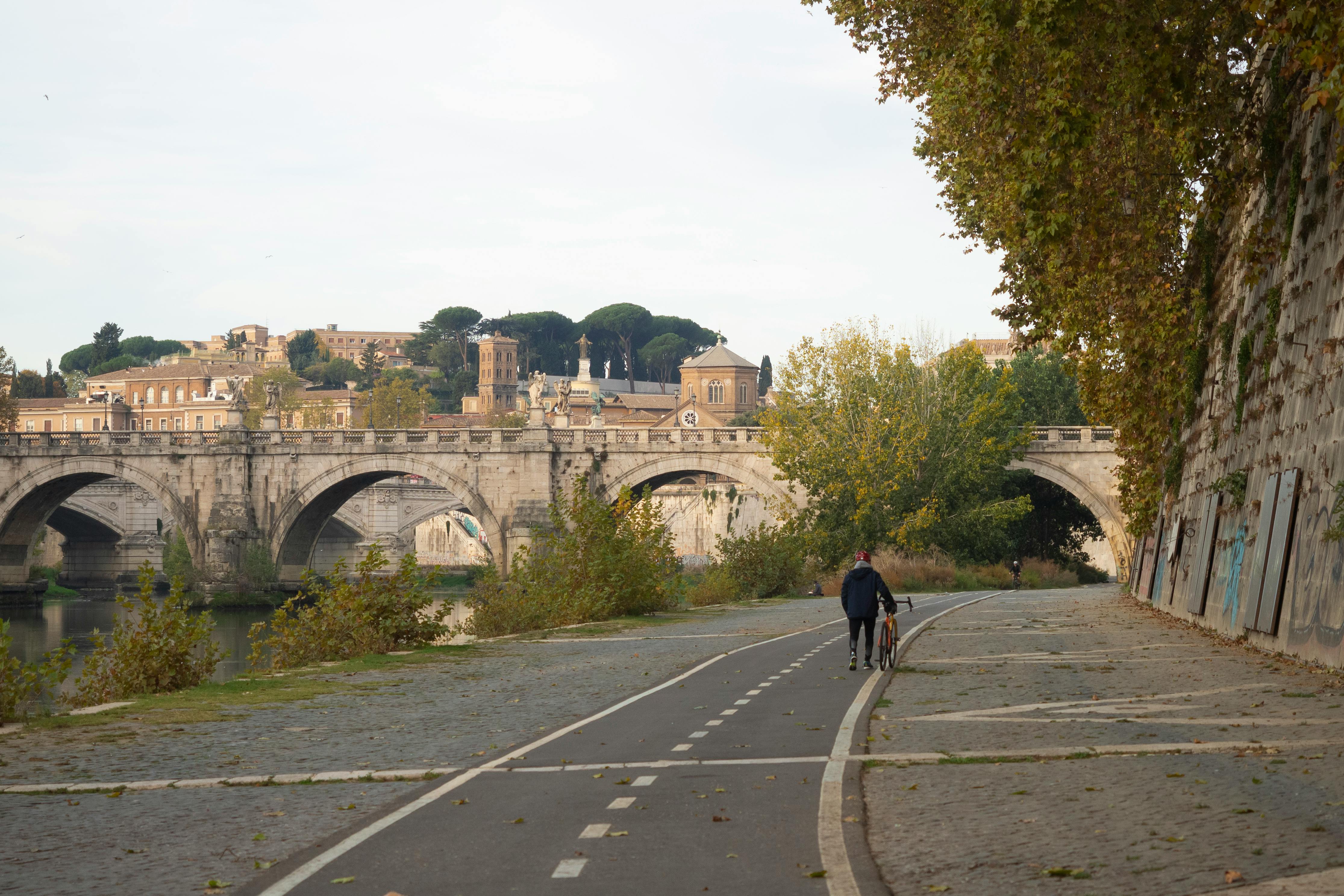 A cyclist walks along a path by a historic bridge in Rome, Italy.