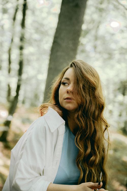 Free A woman with long, wavy hair stands in a sunlit forest, looking contemplative. Stock Photo