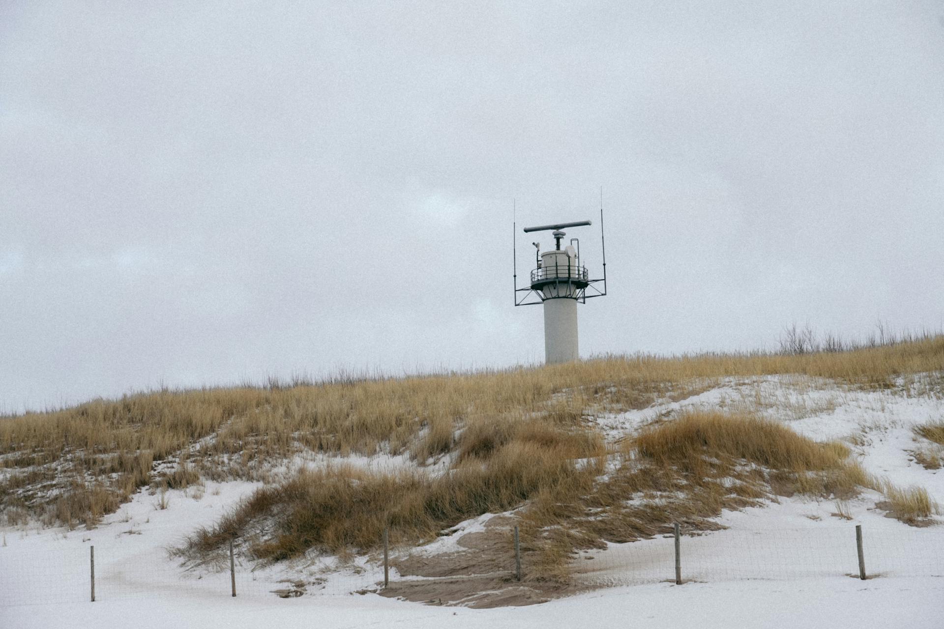 A coastal radar tower stands amidst snow-covered dunes and dry grass under a cloudy winter sky.