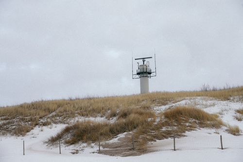 Free A coastal radar tower stands amidst snow-covered dunes and dry grass under a cloudy winter sky. Stock Photo