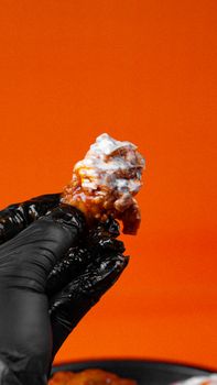 Close-up of a spicy chicken wing held in a black glove against a vibrant orange background.