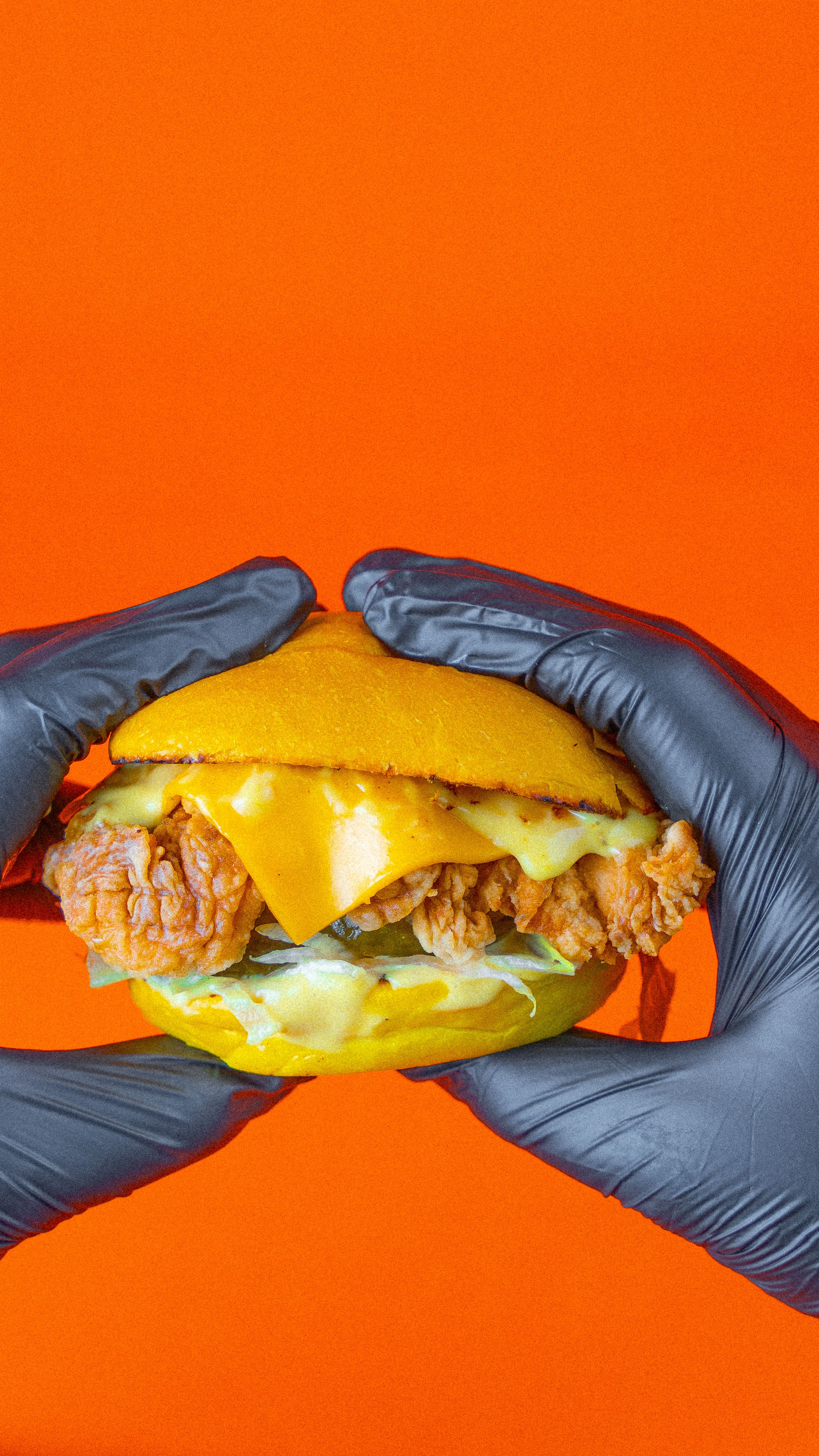 Close-up of a cheeseburger with fried chicken held by hands in black gloves against an orange background.
