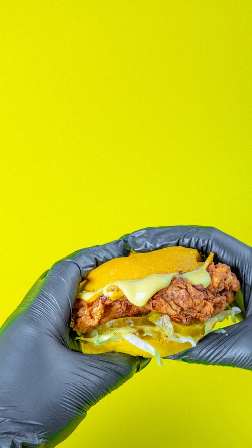 Hands in Black Gloves Holding Chicken Cheeseburger on Bright Yellow Back