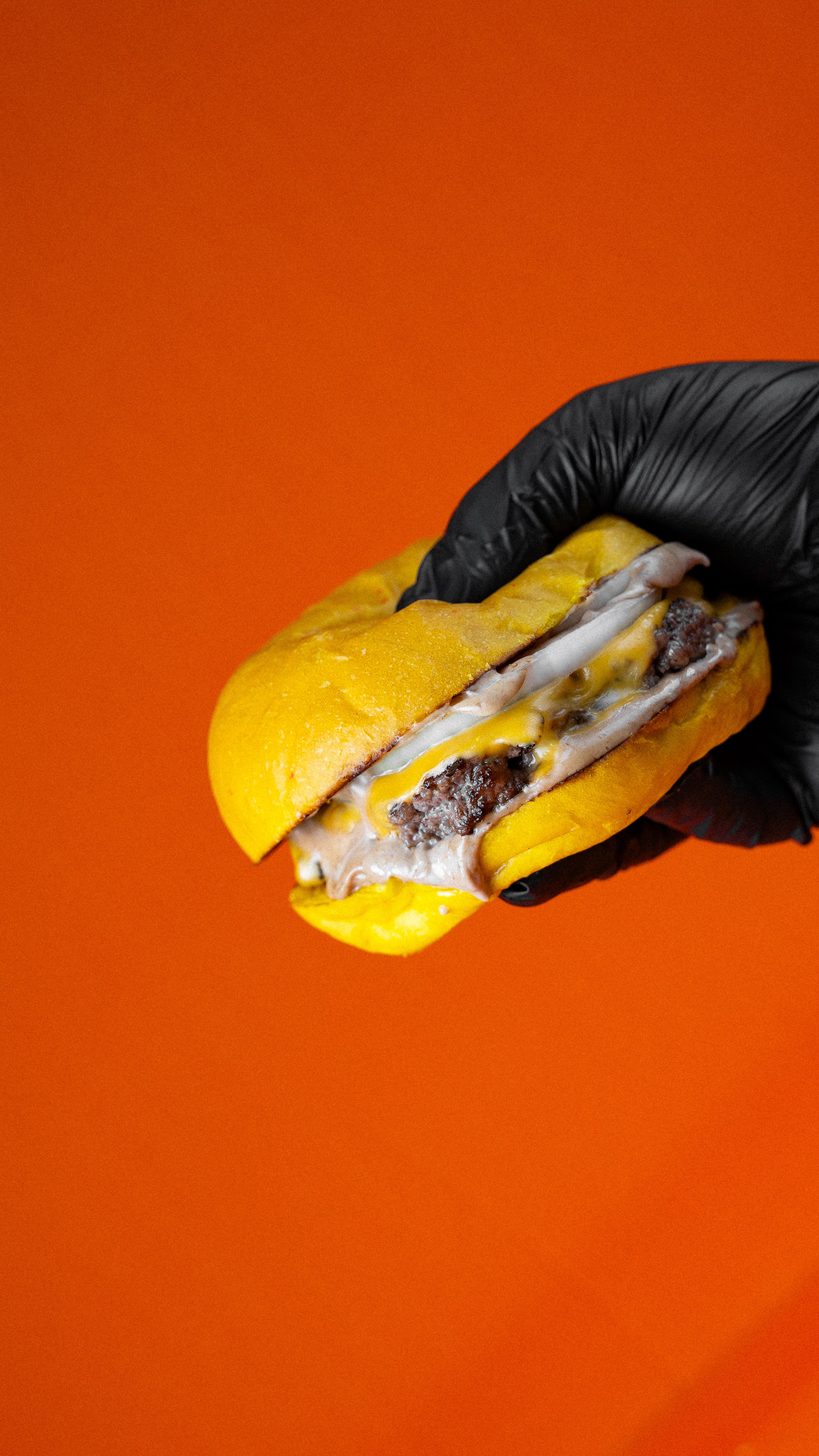 Ali Dashti - Black-gloved hand holding a delicious cheeseburger against a vibrant orange background.