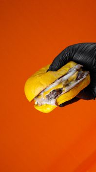 Black-gloved hand holding a delicious cheeseburger against a vibrant orange background.