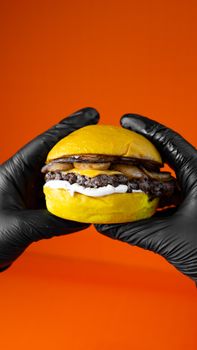Close-up of a juicy cheeseburger held with gloves against an orange background.