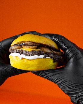 A juicy cheeseburger held by gloved hands with a vibrant orange background.
